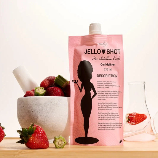 A pink pouch of Jello Shot Strawberry Curl Definer styled on a wooden board with strawberries, a mortar and pestle, and glassware against a clean white background.