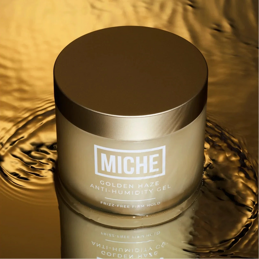 A jar of MICHE Golden Haze Anti-Humidity Gel, featuring a metallic gold lid and frosted glass body, set against a reflective golden liquid background with a clear reflection.