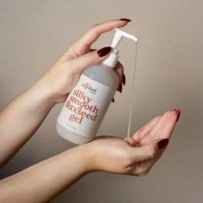 A person's hands with dark red manicured nails are shown holding and dispensing a clear product from a frosted white pump bottle labeled 'miribel silky smooth flaxseed gel' against a solid warm beige background.