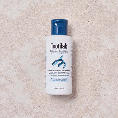 A bottle of Tootilab Detangling Conditioner with a white body and a dark blue cap, placed on a textured beige surface.