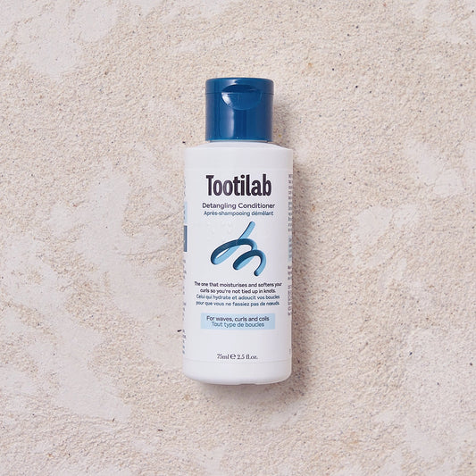 A bottle of Tootilab Detangling Conditioner with a white body and a dark blue cap, placed on a textured beige surface.