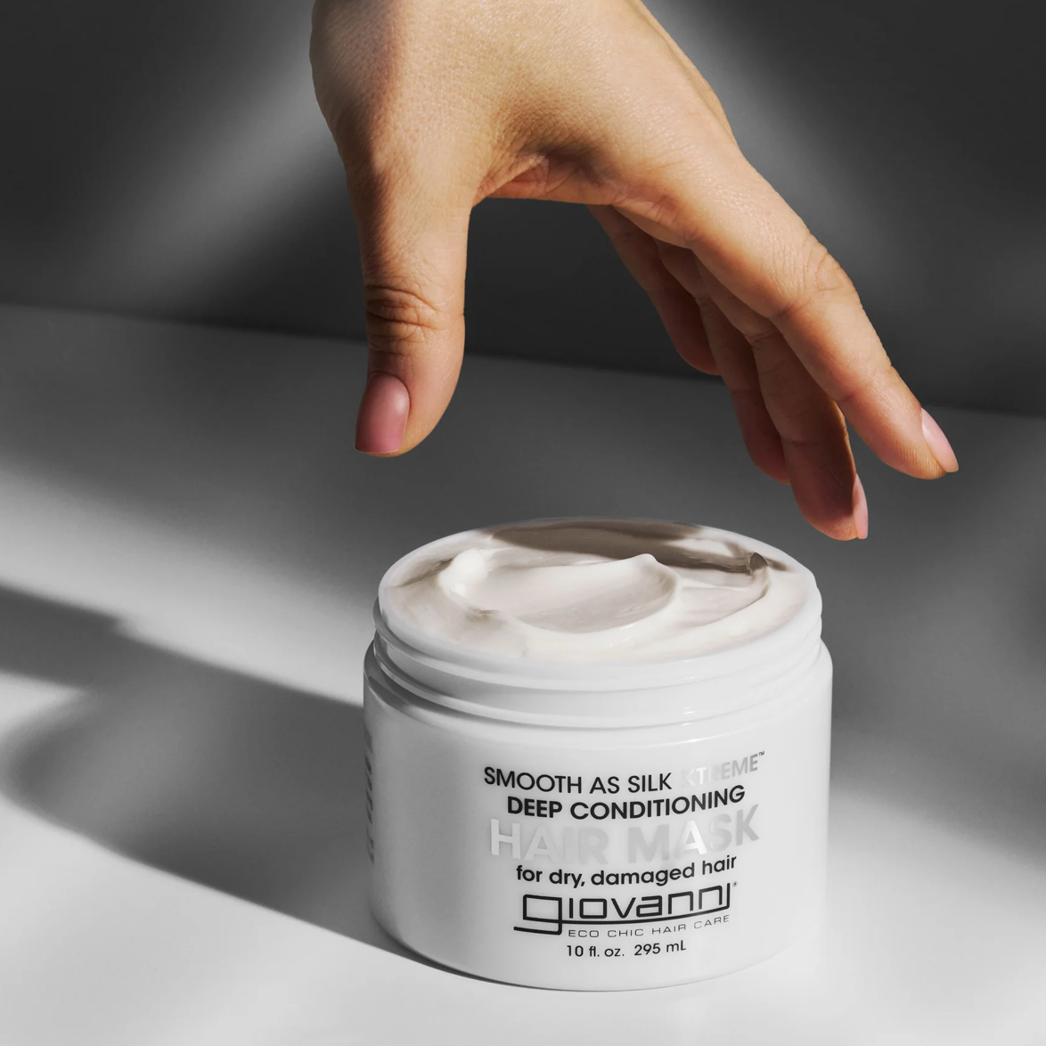 Hand reaching towards a jar of Giovanni hair mask on a neutral background