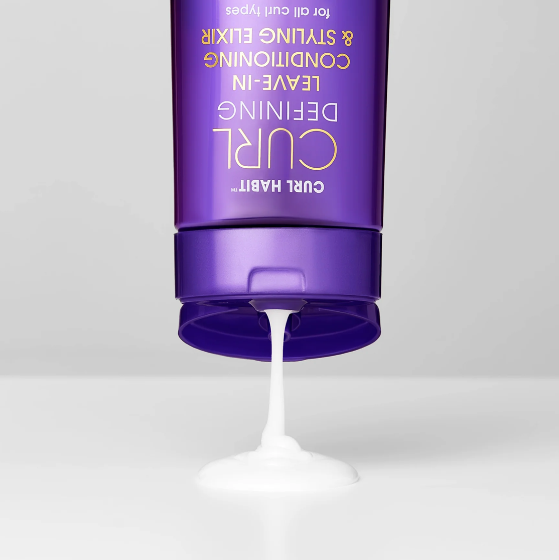 Purple bottle of hair product with white cream being squeezed out on a light gray background