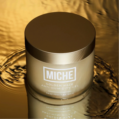 A jar of MICHE Golden Haze Anti-Humidity Gel, featuring a metallic gold lid and frosted glass body, set against a reflective golden liquid background with a clear reflection.