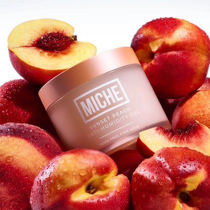 A jar of MICHE Sunset Peach Anti-Humidity Gel surrounded by fresh peach halves and whole peaches on a clean white background.